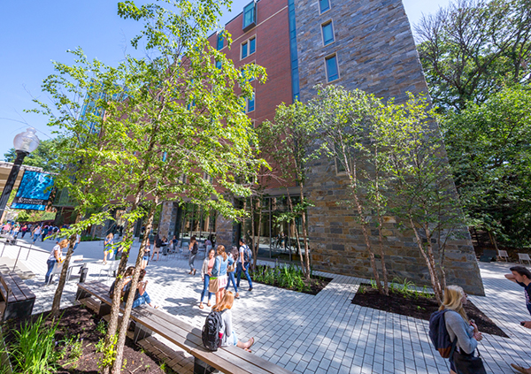 Students walk past a residence hall made of red brick and large glass windows surrouned by green trees and light-colored walkway