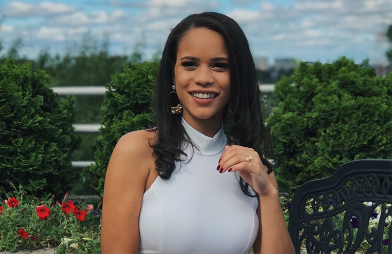 Bianca Uribe smiles outside with trees and red flowers in the background.