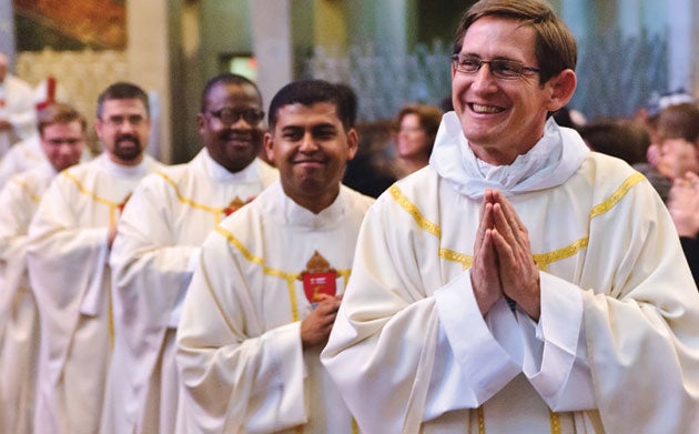 A priest smiles while holding his hands in prayer while other priests process behind him