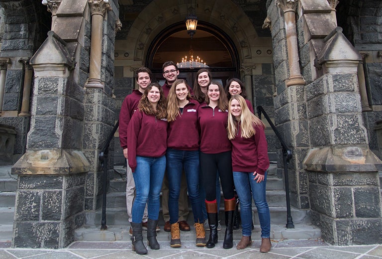 Eight students dressed in maroon shirts stand on the steps of Healy Hall.