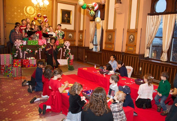 Children kneel at tables decorating while a choir stands above with music folders singing in the Philodemic Room