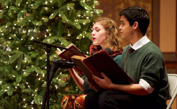 Two singers stand in front of a microphone with their music folders and a tree in the background