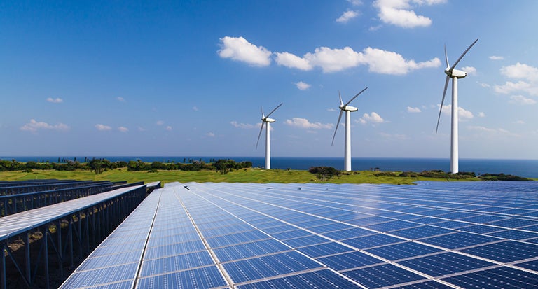 modern wind mills turn with soal panels in the foreground and a blue sky in the distance 