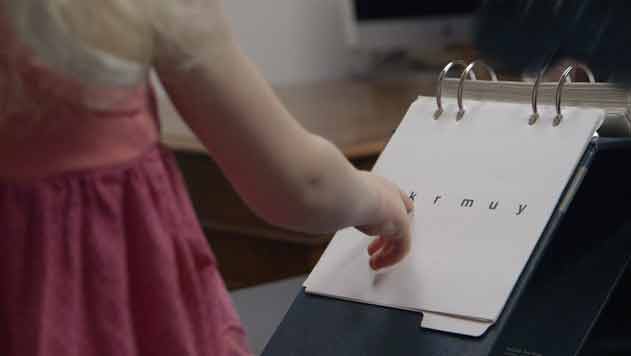 A small child points to a notebook with letters on it during an evaluation of her reading.