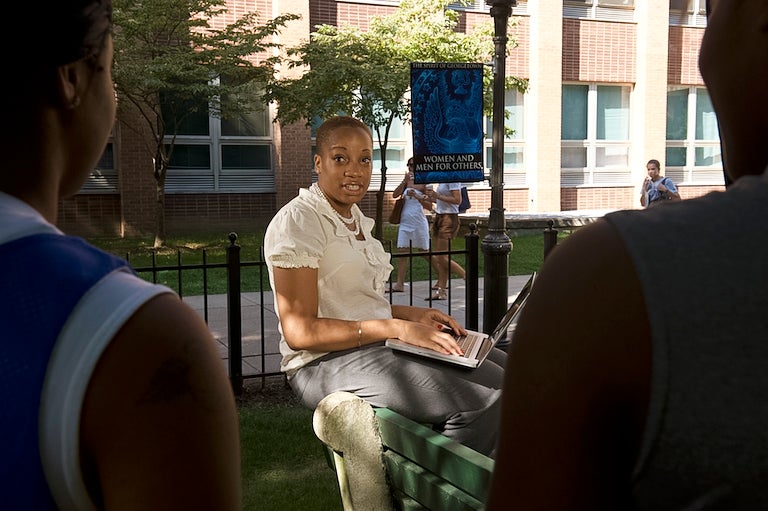 Pamela Nwaoko sits on a stone bench talking while typing with a laptop in her lap.