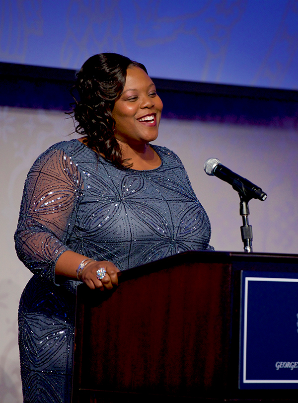 Kaya Henderson speaks from behind a lectern in a sparkling evening dress as she accepts the Halsey Award with a blue backdrop showing in the background.