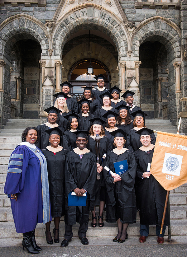Kaya Henderson stands in academic regalia with eighteen recent graduates dressed in their caps and gowns standing in front of Healy Hall
