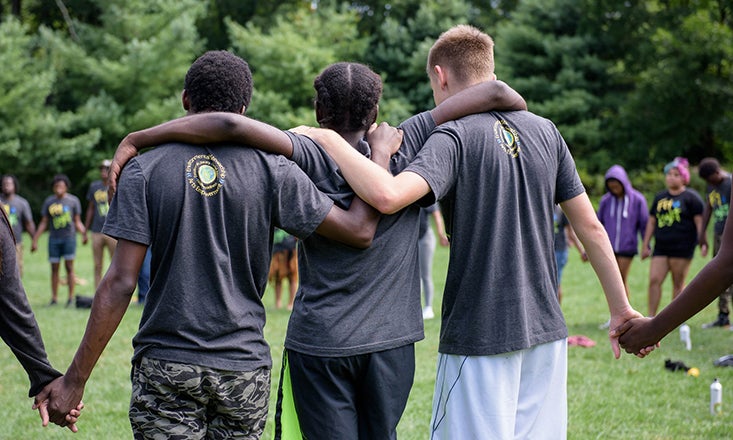 young people with arms draped around one another and others holding hands in a field facing trees