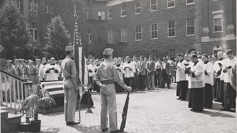 American flag draped over a coffin with another American flag standing near soldiers and priests in Dahlgren Quadrangle at Georgetown  