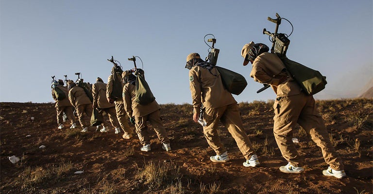 A group of women walk in a straight line carrying equipment through a dry barren region.