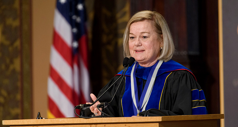 Barbara Bayer speaks at a lectern in Gaston Hall dressed in academic regalia with the American flag in the background.