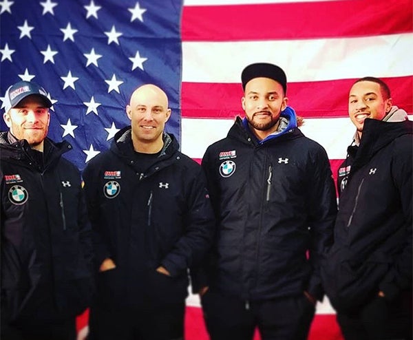 Four Members of the U.S. Olympic Men's Bobsled Team stand in front of the U.S. flag.