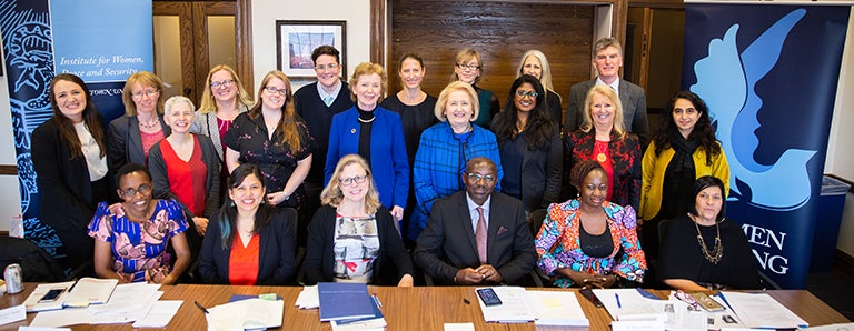 A group of 25 mostly female global leaders and facilitators sit and stand together in a conference room.