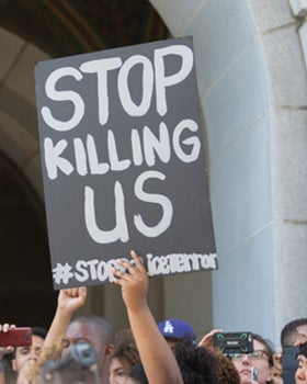 A protestor holds a black sign with the words "Stop Killing Us" written on it.