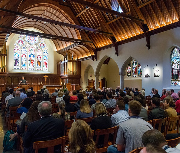 Members of the university community in the foreground sit watching and listening to the archbishop as he stands in front of colorful stained glass under the chapels's wooden arches.