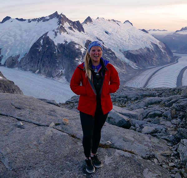 Deirdre Collins smiles while standing in the middle of an icefield in Juneau, Alaska, with snow-capped mountains in the background.