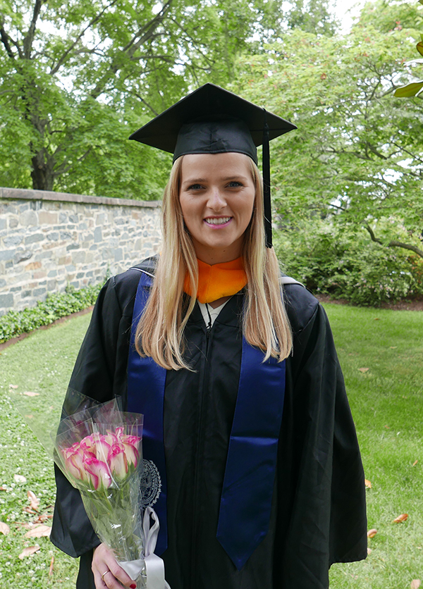 Deirdre Collins stands in her graduation cap and gown holding a bouquet of flowers outside with a background of greenery and stone wall.