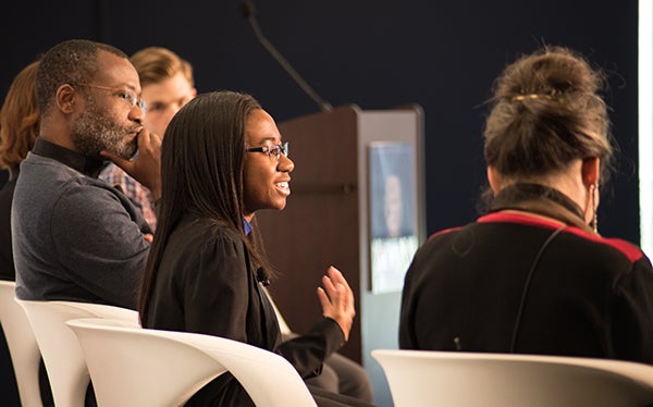 A side profile shot shows Deirdre Jonese Austin speaking during the panel discussion with Rev. Ludovic Lado, S.J., in the background sitting beside her.