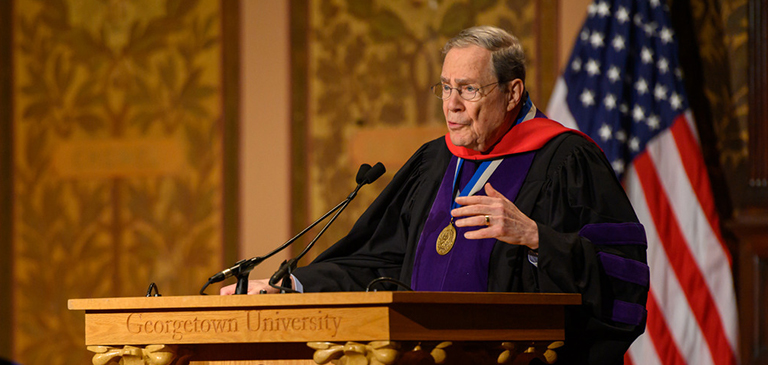 Peter Edelman speaks into the microphone at a lectern while wearing academic regalia in Gaston Hall.