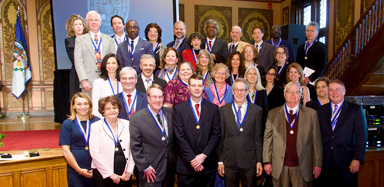 A group of more than 30 faculty members and academic professionals pose on stage in Gaston Hall with gold and silver medals hanging around their necks.