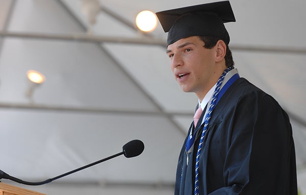 David Fajgenbaum speaking into a microphone at a podium in cap and gown.