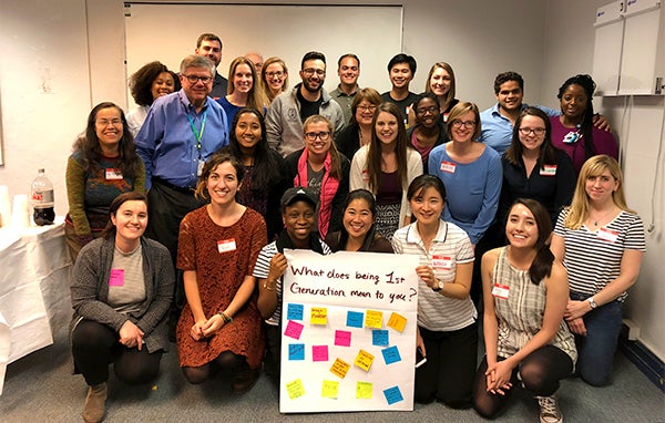 A group of first-generation medical school students pose together holding up a poster that reads "What Does Being 1st Generation Mean to You?"