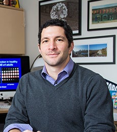 Adam Green sits at his desk with hos work on a computer screen in the background.