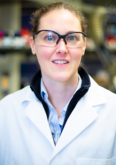 Karah knope stands in her lab dressed in a white coat and goggles.