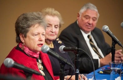 Carol Lancaster speaks with a microphone at a table while Madeleine Albright and Harold White look on