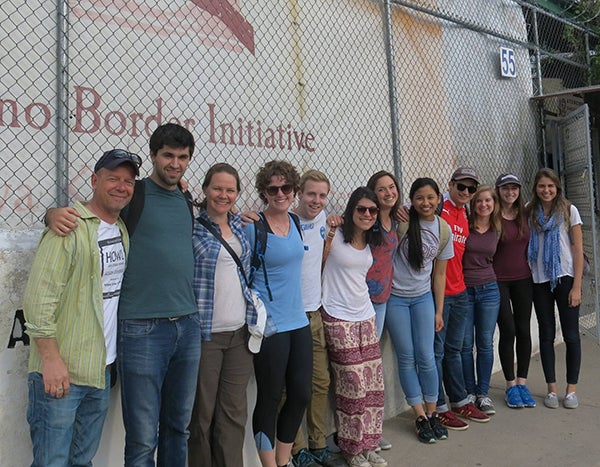 Tomas Alvarez Belon stands with members of the Georgetown community in front of a fence at the Kino border with the Kino Border initiative printed on a wall behind them.