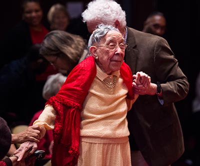 Daisy Pebbles Sewell, 95, stands with assistance from an audience member to cheers and applause.