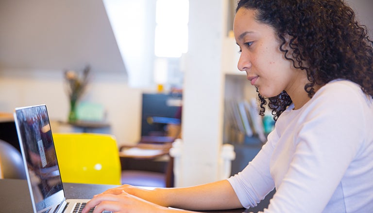 Leslie Telleria sits at a desk while typing on a laptop in a bright room with white walls in the background.