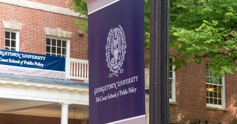 A McCourt School of Public Policy banner hangs on a lampost in front of the red brick Old North building.