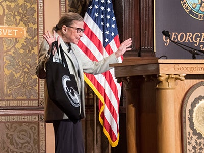 Supreme Ct. Justice Ruth Bader Ginsburg waves to audience holding bag in front of American flag