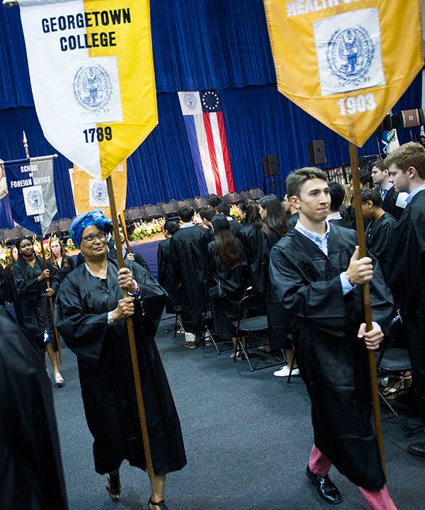 Mélisande Short-Colomb dressed in an academic robe holds the yellow and white Georgetown College Banner on stage with faculty dressed in academic regalia