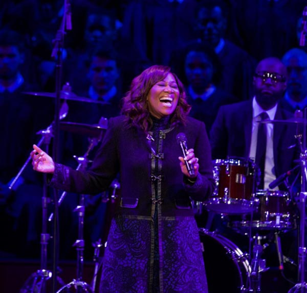 Yolanda Adams sings with microphone while man plays drums in the background with choir
