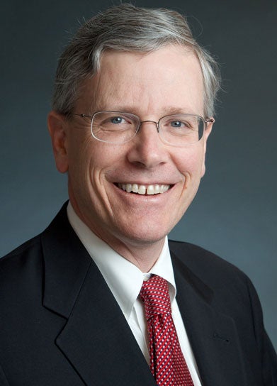 John Monahan, in a suit and tie, smiles for the camera against a gray background. 