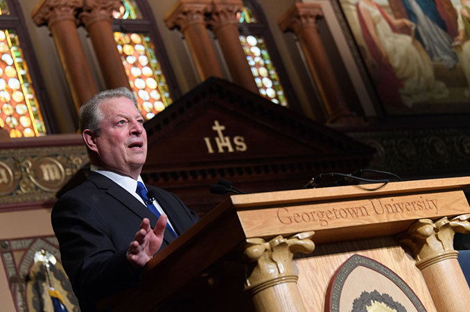 Al Gore speaking at podium in Gaston Hall with stained glass windows behind him