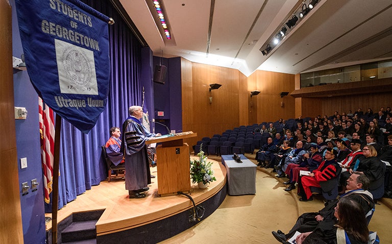 Robert Groves speaks from the stage as he audience looks on and a Georgetown banner appears in the foreground.