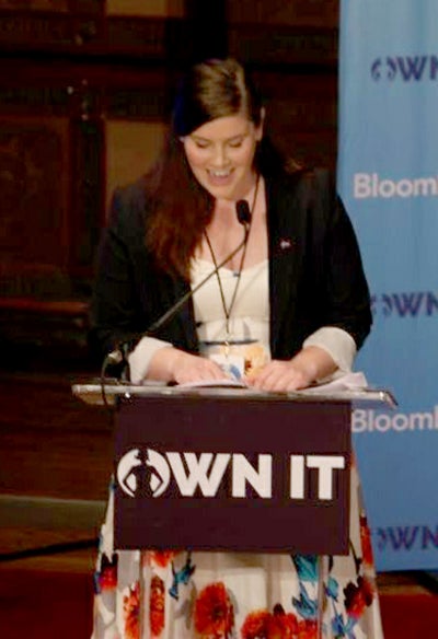 Cristine Pedersen looks down as she speaks at a lectern in Gaston Hall with the OWN IT Summit sign in front.