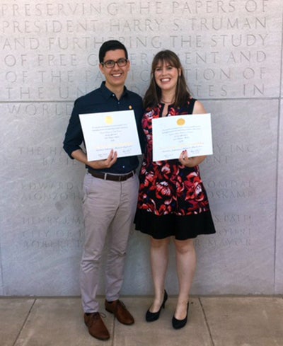 Daniel Wassim and Cristine Pedersen stand together smiling in front of a gray stone building holding their Truman Scholarship certificates.