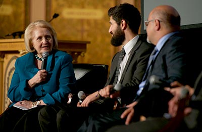 Melanne Verveer speaks while seated and holding a microphone, facing two men during a panel discission. 