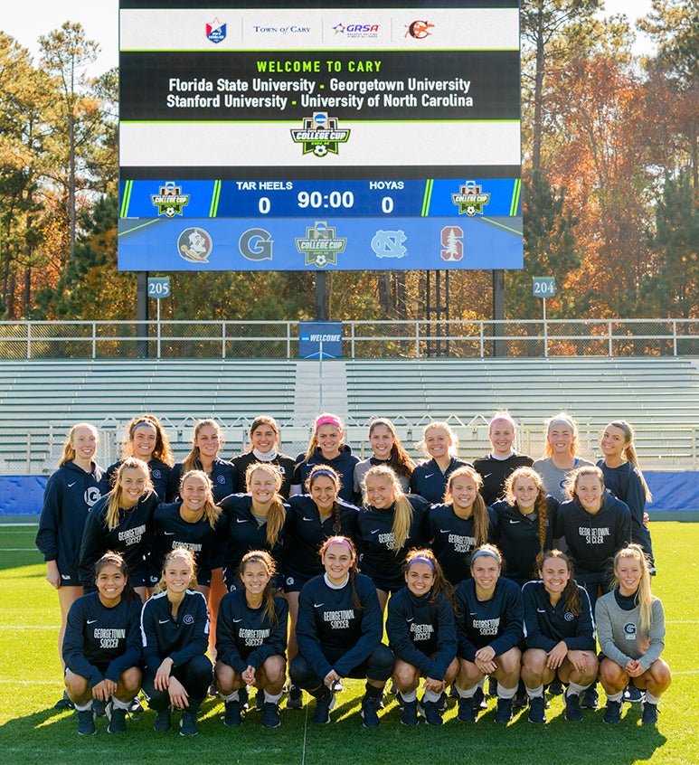 Members of the women's soccer team pose together on the filed in three rows with autumn-leaved trees in the background. 