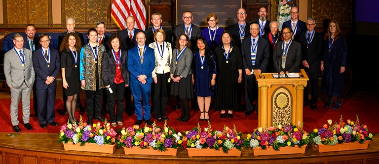 A group of 25 female and male faculty members wear their Vicennial medals on stage in Gaston Hall.