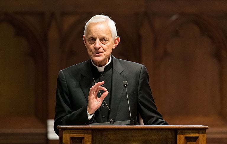 Cardinal Donald Wuerl speaks at a wooden lectern in Dahlgren Chapel with the building's interior woodwork displayed in the background