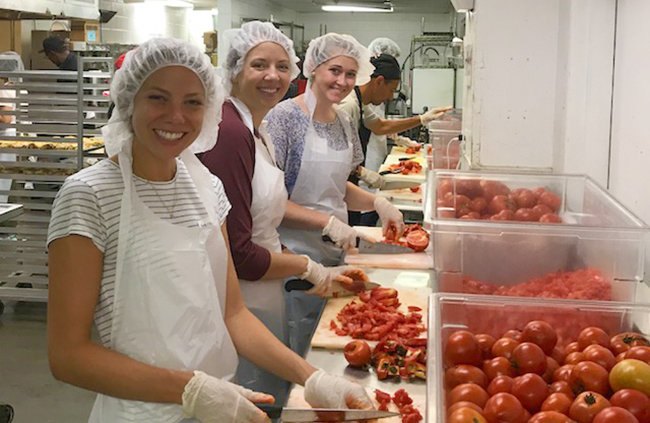 Three female students smile as they chop bright red tomatoes in a professional kitchen.