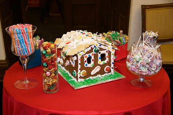 A gingerbread house sits on a table with a red cloth surrounding by glasses of candy