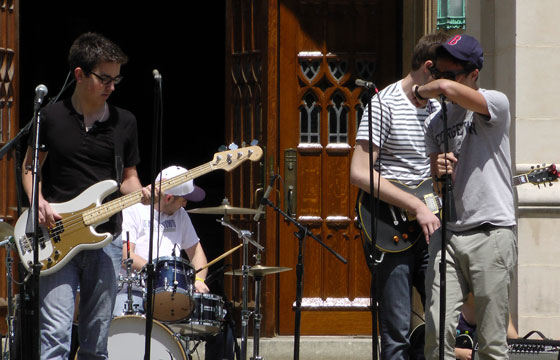 A student sings into a microphone while others play a bass, guitar and drums behind him on White Gravenor Patio