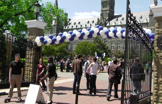 People congregate underneath a white and blue balloon arch across the Healy Gates with Healy Hall in the background