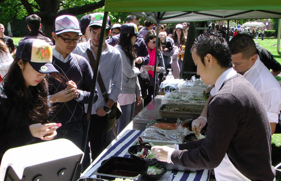 Students wait in line to be served food underneath a tent from trays